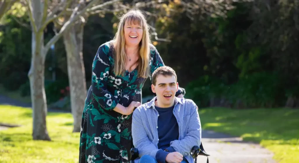 Two people smiling at the camera in the sunshine, one in a wheelchair