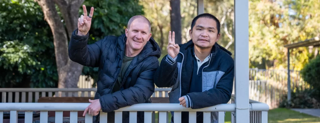 Two men smiling at the camera leaning on a railing, both giving the peace sign