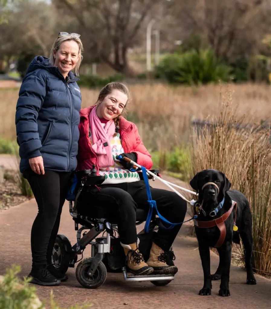 Two women and their guide dog on a walk. One is in a wheelchair and the dog is in it's harness