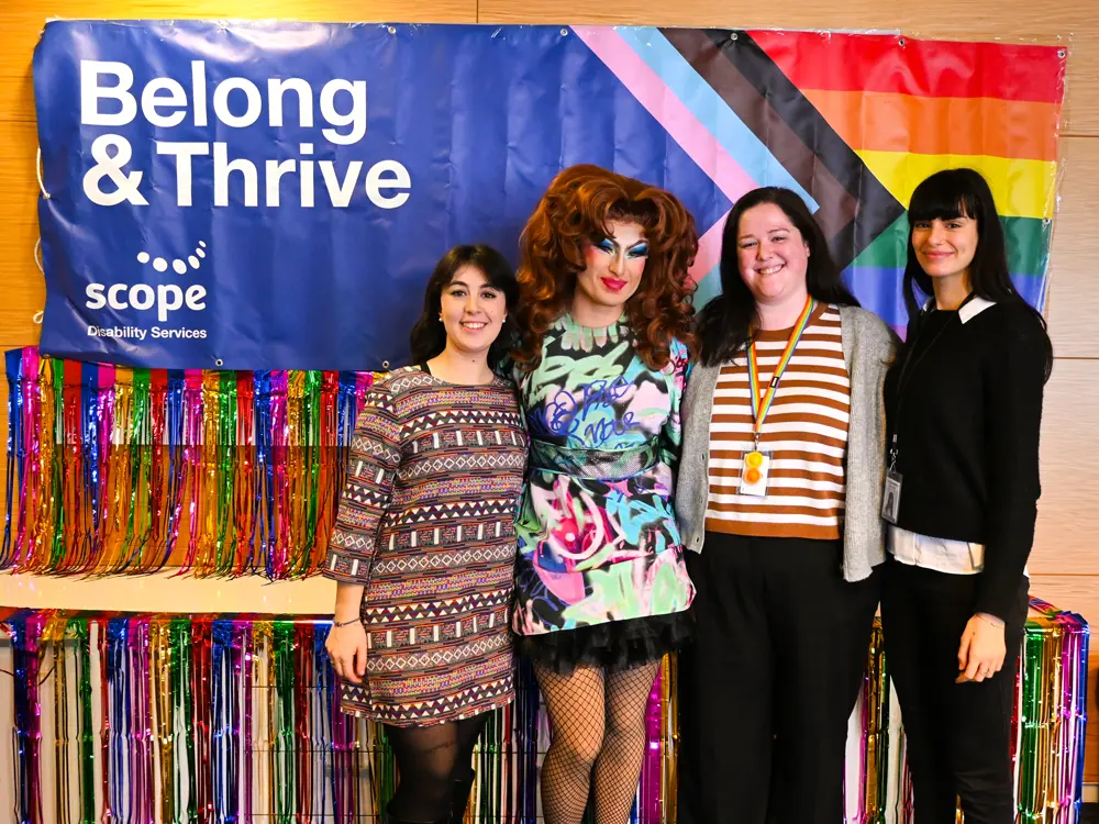 Belong and Thrive - Pride week at scope, a group of women with a drag queen infront of the pride flag and rainbow tinsel