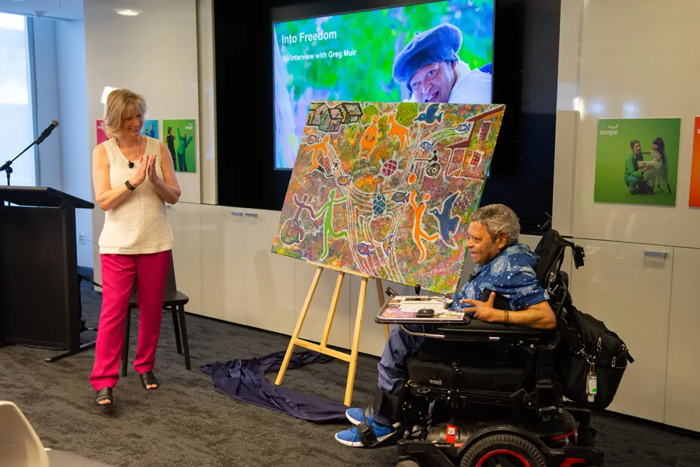 Woman standing up and clapping, next to an artwork completed by Greg Muir, with the artist next to the artwork in his wheelchair. The image in the background says "Into Freedom - An interview with Greg Muir"