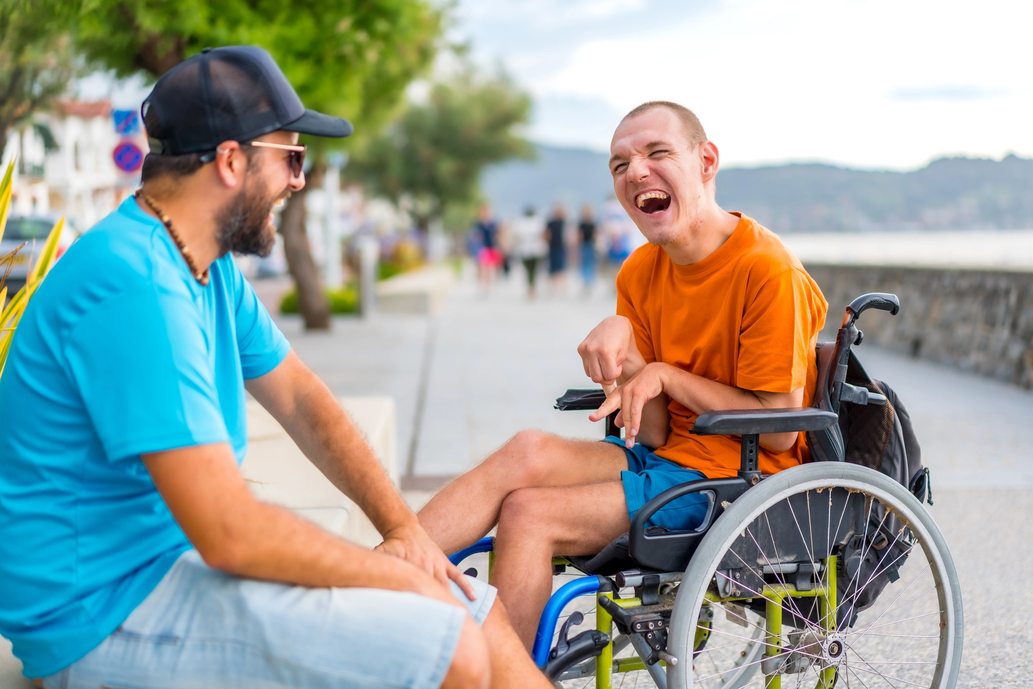 Two people smiling at each other out by the water, one man is in a wheelchair and the other is wearing a black cap